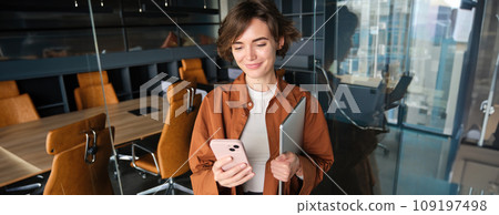 Image of stylish working woman checking her messages on smartphone, holding laptop, standing in an office, working in co-working space Image of stylish working woman checking her messages on smartphone, holding laptop, standing in an office, working in co-working space 109197498
