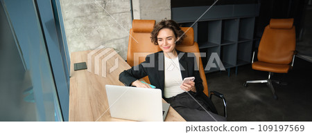 Portrait of young professional, businesswoman sitting in her office, answering phone call, using her smartphone and laptop, working near window 109197569