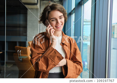 Happy young office worker near window, calling someone, answers a call on mobile phone, talking and smiling 109197767