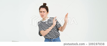 Image of woman expressing aversion, dislike something, looks apalled, extends hands forward in defensive gesture, rejecting, refusing, standing over white background Image of woman expressing aversion, dislike something, looks apalled, extends hands forward in defensive gesture, rejecting, refusing, standing over white background 109197838