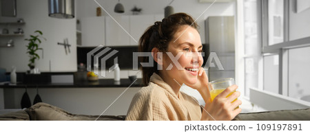 Close up of beautiful young woman on sofa, drinks orange juice and smiles, looks outside window, enjoys morning at home 109197891