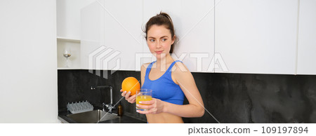 Indoor shot of young brunette woman in sportswear, drinking orange juice, holding fruit in hand, posing in kitchen Indoor shot of young brunette woman in sportswear, drinking orange juice, holding fruit in hand, posing in kitchen 109197894