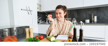 Portrait of beautiful smiling woman, writing her healthy menu, eating tomato while cooking, making grocery list, sitting in the kitchen 109197940