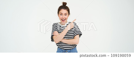 Image of excited young woman, smiling, showing amazing price discounts, pointing left, demonstrating something awesome, standing over white background 109198059