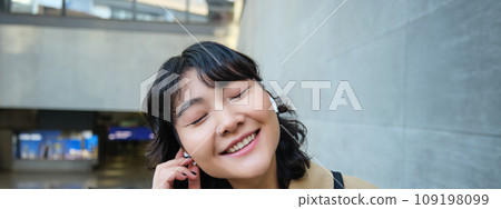 Close up portrait of smiling korean girl in headphones, listens music in wireless earphones, enjoys travelling around town with her favorite songs playlist 109198099