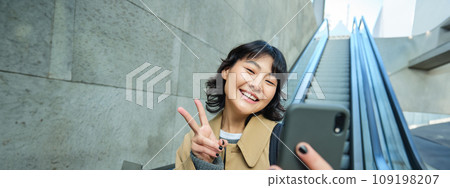 Positive asian girl in trench, shows peace sign, takes selfie while goes down escalator, smiles and looks happy Positive asian girl in trench, shows peace sign, takes selfie while goes down escalator, smiles and looks happy 109198207