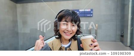 Portrait of stylish asian woman in headphones, drinks coffee to go and smiles, enjoys cappuccino while commutes, stands on street Portrait of stylish asian woman in headphones, drinks coffee to go and smiles, enjoys cappuccino while commutes, stands on street 109198309