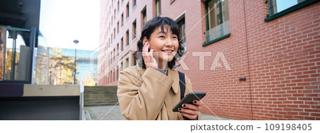 Smiling brunette woman, asian student walks on street and listens music in wireless earphones, holds smartphone, reads text message on mobile phone, picks song from playlist Smiling brunette woman, asian student walks on street and listens music in wireless earphones, holds smartphone, reads text message on mobile phone, picks song from playlist 109198405