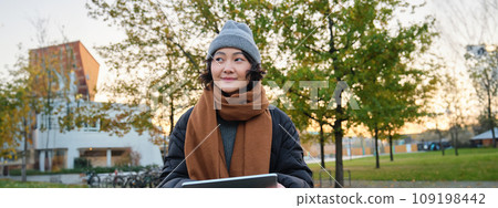 Smiling young girl, korean student sits in park in scarf and hat on chilly day, draws outdoors on digital tablet with graphic pen, makes scatches Smiling young girl, korean student sits in park in scarf and hat on chilly day, draws outdoors on digital tablet with graphic pen, makes scatches 109198442