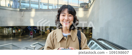 People in city. Young korean woman travels around city, goes up the escalator, uses her mobile phone and smiles People in city. Young korean woman travels around city, goes up the escalator, uses her mobile phone and smiles 109198607
