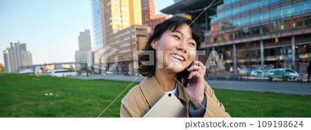University lifestyle. Young asian student, girl with tablet and notebook, talks on mobile phone while walks from campus on street University lifestyle. Young asian student, girl with tablet and notebook, talks on mobile phone while walks from campus on street 109198624