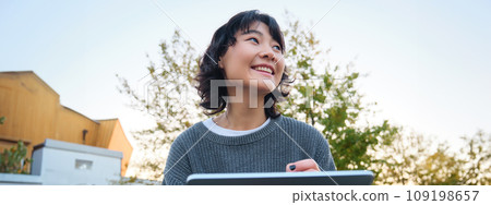 Close up portrait of young korean girl sits outdoors in park, holds her digital tablet and graphic pen, draws scatches, gets inspiration from nature for art, smiles happily 109198657