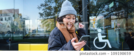 Modern people and lifestyle. Happy asian girl screams from joy, celebrates, stands near bus public transport and looks amazed Modern people and lifestyle. Happy asian girl screams from joy, celebrates, stands near bus public transport and looks amazed 109198659