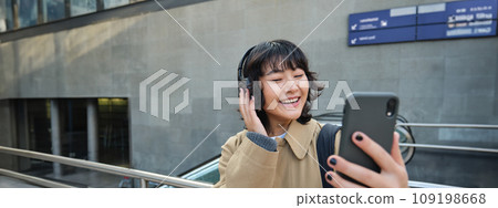Beautiful korean girl in headphones, takes selfie on her smartphone, stands on street with mobile phone, records video, listens music and travels around city Beautiful korean girl in headphones, takes selfie on her smartphone, stands on street with mobile phone, records video, listens music and travels around city 109198668