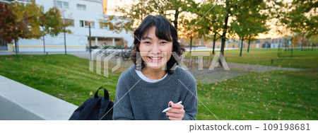 Young asian girl with graphic pencil and tablet, sits in park on bench, draws scatches, does her homework outdoors Young asian girl with graphic pencil and tablet, sits in park on bench, draws scatches, does her homework outdoors 109198681