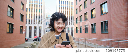 Portrait of cute girl tourist, korean woman in headphones, looks at mobile phone, uses smartphone app, map or text messages, listens music in headphones Portrait of cute girl tourist, korean woman in headphones, looks at mobile phone, uses smartphone app, map or text messages, listens music in headphones 109198775