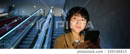 Close up portrait of smiling asian brunette girl, going down escalator, holding smartphone and listen music in headphones, using public transport, travelling in city 109198815