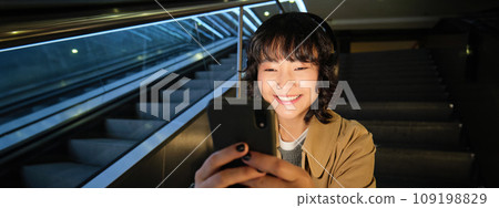 Portrait of happy asian girl in headphones, smiling and looking at smartphone, watching video or listening music on mobile phone, sitting on staircase 109198829