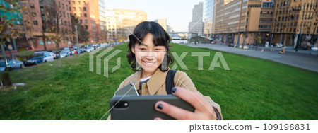 Happy young woman, student takes selfie on street, holds her tablet and homework notes, uses smartphone 109198831