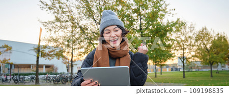 Portrait of happy asian girl sits on bench, looks at digital tablet screen and cheers, triumphs, wins something, celebrates good news, relaxes in park outdoors Portrait of happy asian girl sits on bench, looks at digital tablet screen and cheers, triumphs, wins something, celebrates good news, relaxes in park outdoors 109198835