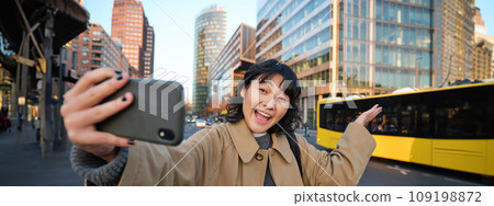 Upbeat asian girl takes selfie with smartphone in city centre, showing something behind her with smiling face. Tourist goes sightseeing 109198872