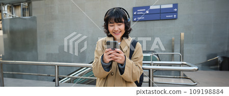 Smiling asian girl in headphones, stands on train station, looks at mobile phone screen and looks pleased, listens music and drinks coffee Smiling asian girl in headphones, stands on train station, looks at mobile phone screen and looks pleased, listens music and drinks coffee 109198884