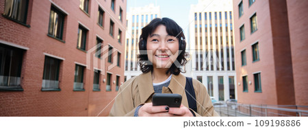 Portrait of cute girl tourist, korean woman in headphones, looks at mobile phone, uses smartphone app, map or text messages, listens music in headphones Portrait of cute girl tourist, korean woman in headphones, looks at mobile phone, uses smartphone app, map or text messages, listens music in headphones 109198886