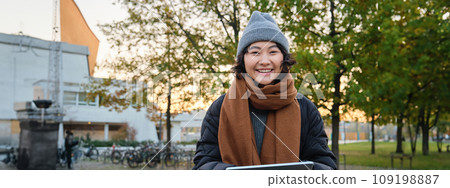 Portrait of asian girl relaxing in park, watching videos or reading on digital tablet, sitting on bench in hat and scarf on chilly day, enjoing outdoors Portrait of asian girl relaxing in park, watching videos or reading on digital tablet, sitting on bench in hat and scarf on chilly day, enjoing outdoors 109198887