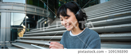 Young woman draws on her tablet, listens to music in headphones. Asian girl sits on stairs and does graphic design project, sits on street stairs 109198890