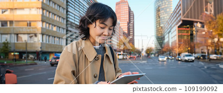 Woman walking on street using digital tablet, holding gadget in hands and looking away, standing in city centre Woman walking on street using digital tablet, holding gadget in hands and looking away, standing in city centre 109199092