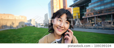 University lifestyle. Young asian student, girl with tablet and notebook, talks on mobile phone while walks from campus on street University lifestyle. Young asian student, girl with tablet and notebook, talks on mobile phone while walks from campus on street 109199104