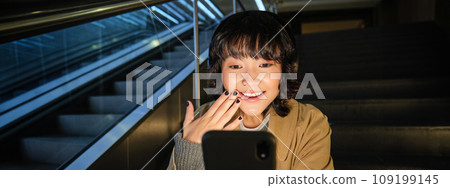 Korean girl looks surprised and happy at her phone screen, smiles amazed, reads good news, sits on staircase near escalator in mall, listens music in headphones Korean girl looks surprised and happy at her phone screen, smiles amazed, reads good news, sits on staircase near escalator in mall, listens music in headphones 109199145