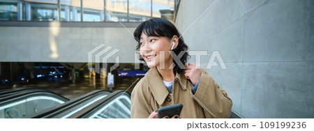 Young brunette woman commutes, goes somewhere in city, stands on escalator and uses mobile phone, holds smartphone and smiles, listens music in wireless headphones Young brunette woman commutes, goes somewhere in city, stands on escalator and uses mobile phone, holds smartphone and smiles, listens music in wireless headphones 109199236