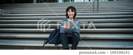 Young woman draws on her tablet, listens to music in headphones. Asian girl sits on stairs and does graphic design project, sits on street stairs Young woman draws on her tablet, listens to music in headphones. Asian girl sits on stairs and does graphic design project, sits on street stairs 109199241