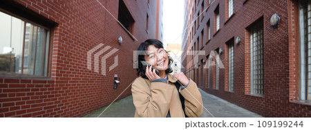 Portrait of young korean woman walking down street with phone, talking with someone, makes a call, has telephone conversation Portrait of young korean woman walking down street with phone, talking with someone, makes a call, has telephone conversation 109199244