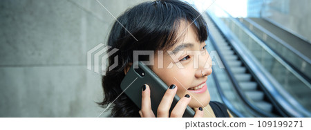 Headshot of smiling korean woman with smartphone, makes a phone call, goes down escalator in city, commutes to university Headshot of smiling korean woman with smartphone, makes a phone call, goes down escalator in city, commutes to university 109199271