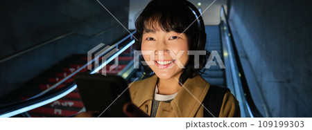 Close up of cute asian girl in headphones, picks song to listen while commuting, going down escalator to metro, smiling as reading message on mobile phone Close up of cute asian girl in headphones, picks song to listen while commuting, going down escalator to metro, smiling as reading message on mobile phone 109199303