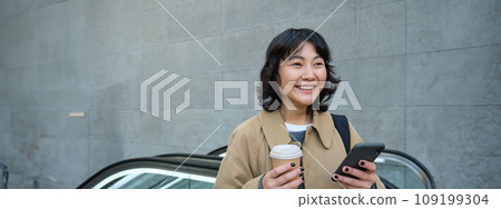 Beautiful happy asian girl, drinks coffee to go, using mobile phone while standing on escalator, walking in city centre and smiling 109199304
