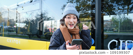 Enthusiastic girl rejoice, reads message on mobile phone and celebrates, stands near her bus on public transport stop and looks excited, posing in warm winter clothes Enthusiastic girl rejoice, reads message on mobile phone and celebrates, stands near her bus on public transport stop and looks excited, posing in warm winter clothes 109199307