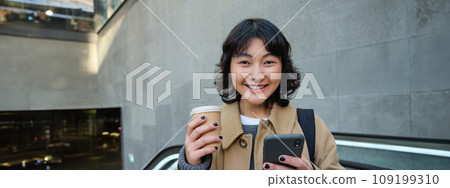 Portrait of hipster girl on escalator, drinking coffee and looking at smartphone, going to work, commute in city, smiling at camera 109199310