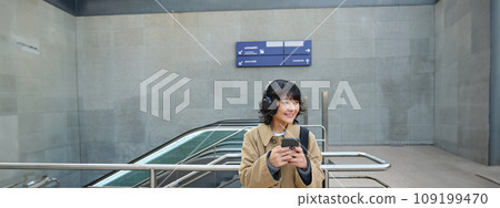 Portrait of stylish asian hipster girl, listens music in headphones, looks at her phone, tourist looking at map for a way to sightseeing, drinking coffee to go Portrait of stylish asian hipster girl, listens music in headphones, looks at her phone, tourist looking at map for a way to sightseeing, drinking coffee to go 109199470
