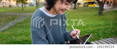 Portrait of young smiling korean girl, graphic designer, artist drawing on digital tablet with a pen tool, sitting in park on fresh air and scatching, taking notes Portrait of young smiling korean girl, graphic designer, artist drawing on digital tablet with a pen tool, sitting in park on fresh air and scatching, taking notes 109199534