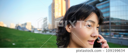 Headshot of young korean woman walks on street, talks on mobile phone, has telephone conversation, stands near campus building Headshot of young korean woman walks on street, talks on mobile phone, has telephone conversation, stands near campus building 109199634