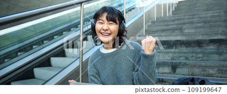 Cheerful korean girl in headphones, sits on stairs with laptop, celebrates, say yes, triumphs, receives good news, success on working project, works remotely on street 109199647