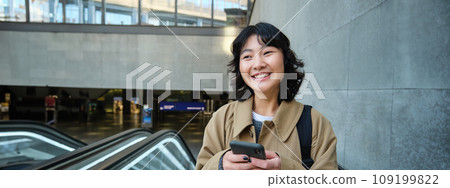 Portrait of cute korean girl in trench coat, going up escalator, holds mobile phone and smiles pleased, commutes 109199822