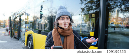 Portrait of confused young woman, looking at her smartphone app with shocked and disappointed face, shrugs shoulders, reads bad news, stands on bus stop near public transport 109199833