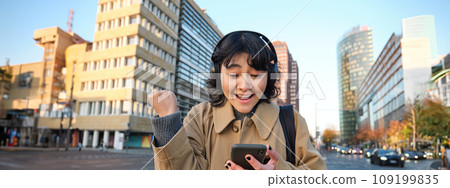 Young happy woman celebrating on street, holding smartphone and cheering, reacts amazed to good news, reads phone text message with surprised joyful face 109199835