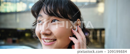 Close up portrait of smiling korean girl in headphones, listens music in wireless earphones, enjoys travelling around town with her favorite songs playlist 109199884