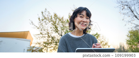 Close up portrait of young korean girl sits outdoors in park, holds her digital tablet and graphic pen, draws scatches, gets inspiration from nature for art, smiles happily Close up portrait of young korean girl sits outdoors in park, holds her digital tablet and graphic pen, draws scatches, gets inspiration from nature for art, smiles happily 109199903