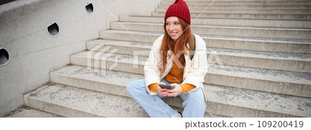 Hipster ginger girl, redhead woman sits on stairs with smartphone, waits for someone and messages on social media on mobile phone app 109200419